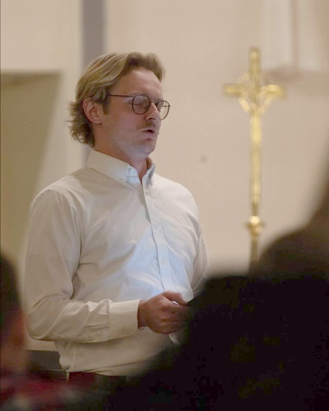Blake Wayment singing in church, a gilt crucifix behind him